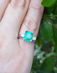 Emerald and side diamonds ring on a finger with a green leafy background.