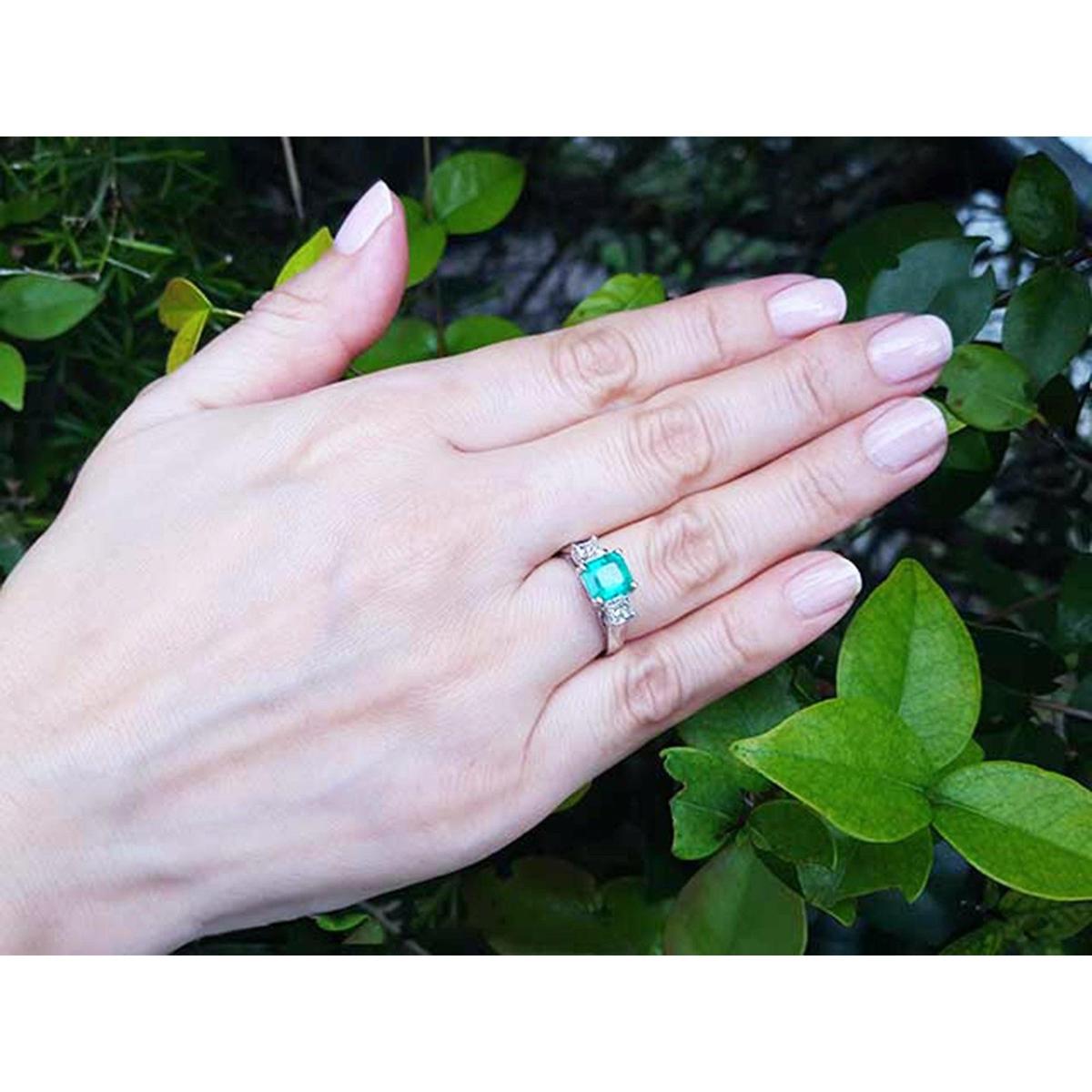 Hand wearing a ring with an emerald stone against a green leafy background.