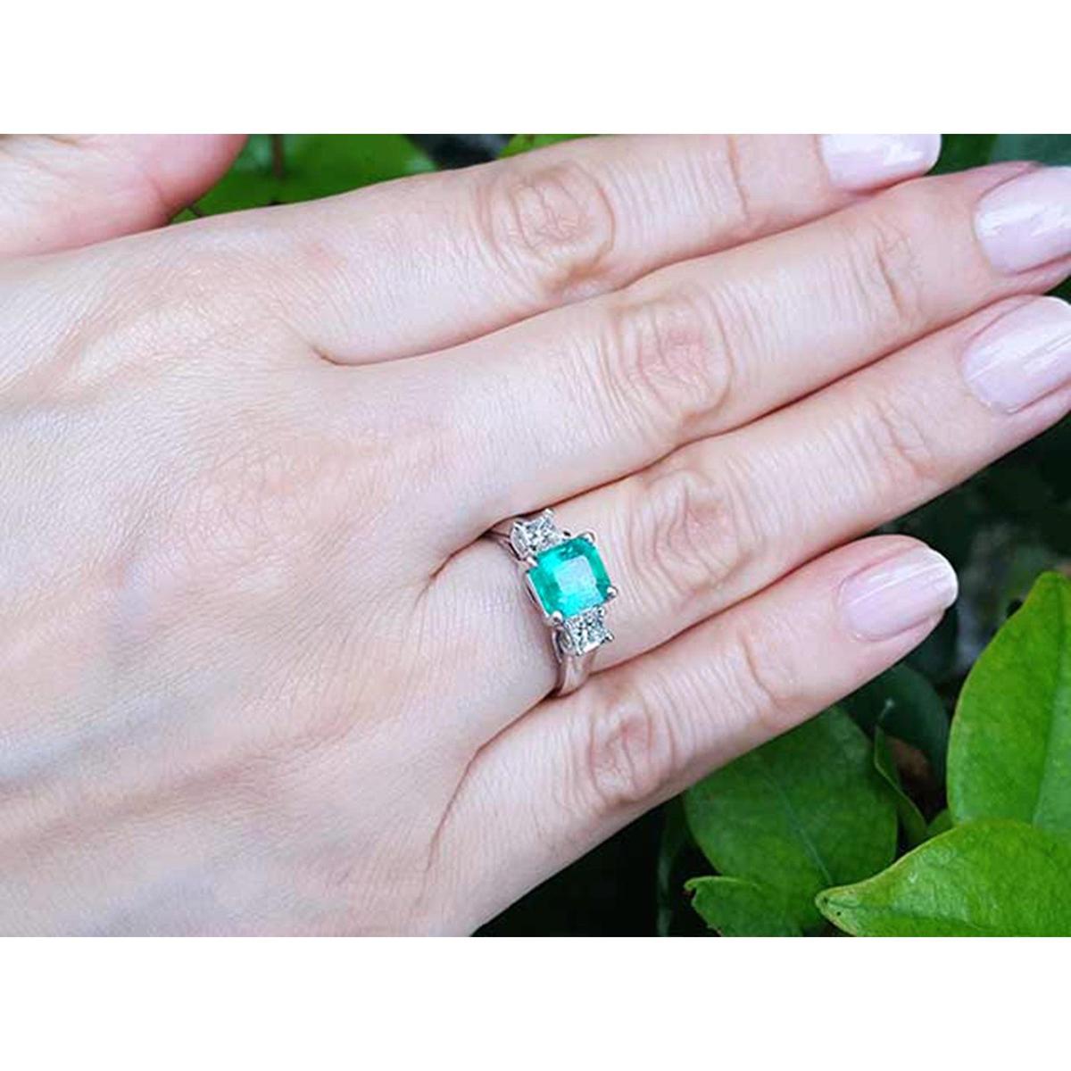 Hand wearing an emerald ring with side diamonds against a green leafy background.