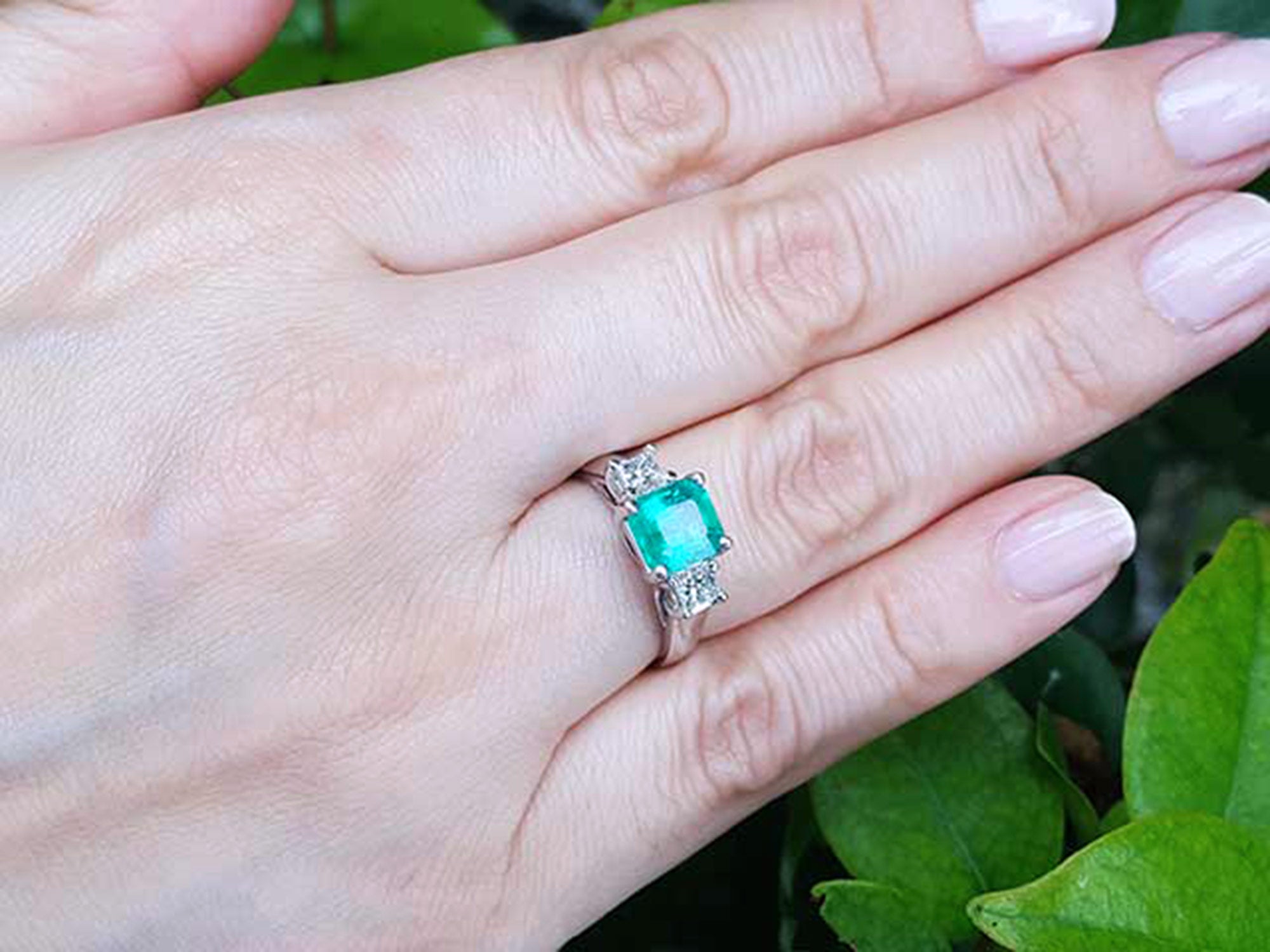Hand wearing an emerald ring with side diamonds against a green leafy background.