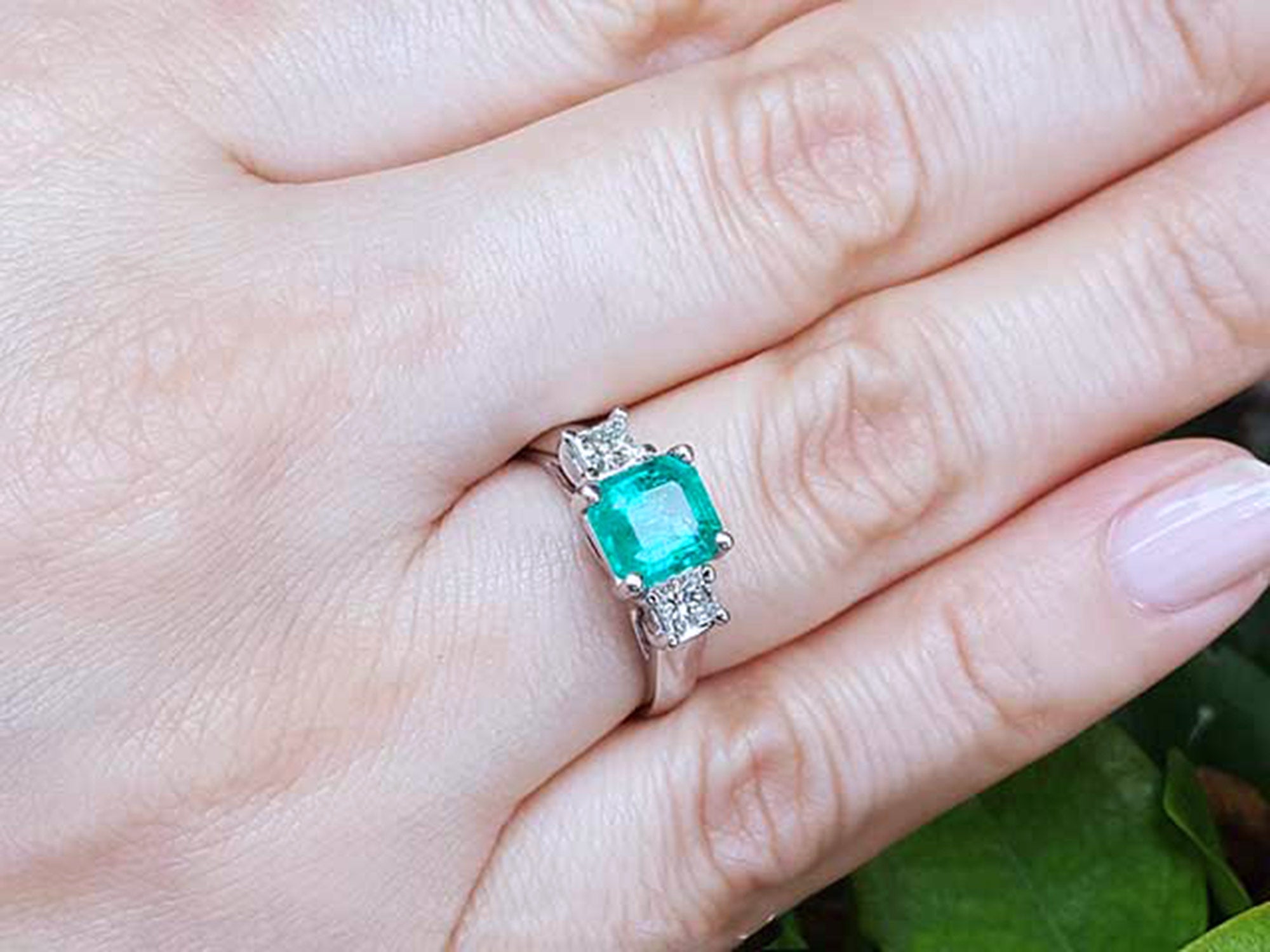 Emerald ring with diamond accents on a hand against a natural background.