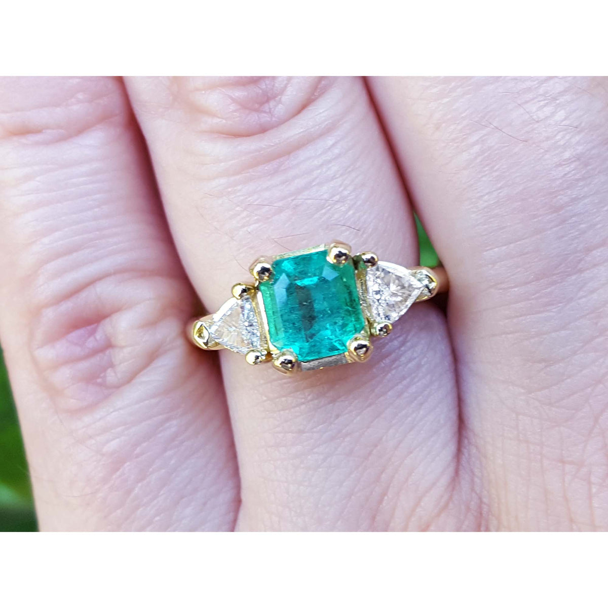 A 18k yellow gold colombian emerald ring with side trillion cut natural diamonds on a finger.