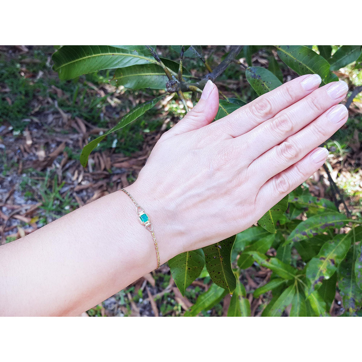A lady&#39;s hand wearing a gold bracelet with a colombian emerald against a natural background.