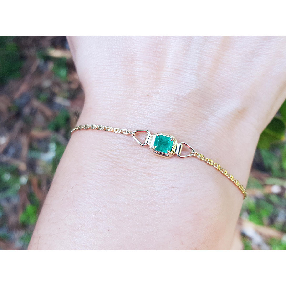 Gold bracelet with a solitaire emerald on a wrist against a blurred natural background.