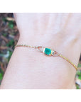 Gold bracelet with a solitaire emerald on a wrist against a blurred natural background.
