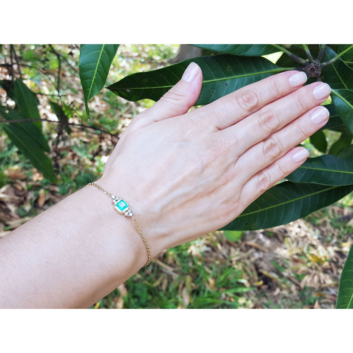 A woman&#39;s hand wearing a gold bracelet with a colombian emerald against a natural background.
