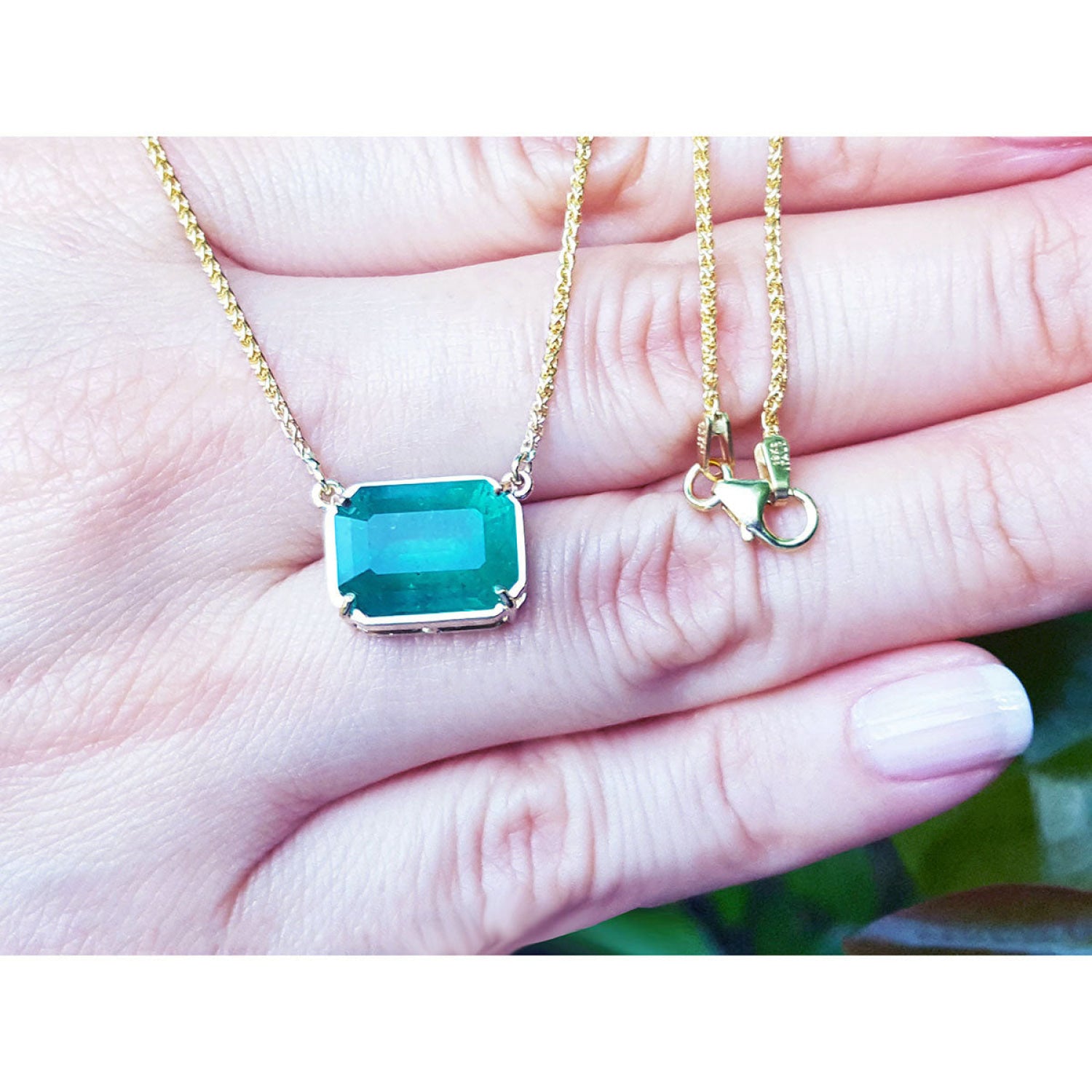 Emerald-cut Colombian emerald solitaire necklace in 18K yellow gold, held in a person's hand to show scale. The necklace chain is gold, and the clasp is visible on the right side of the hand.