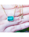 Emerald-cut Colombian emerald solitaire necklace in 18K yellow gold, held in a person's hand to show scale. The necklace chain is gold, and the clasp is visible on the right side of the hand.