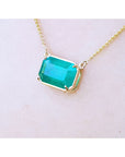 Emerald-cut Colombian emerald solitaire necklace in 18K yellow gold, displayed on a light skin-toned surface.