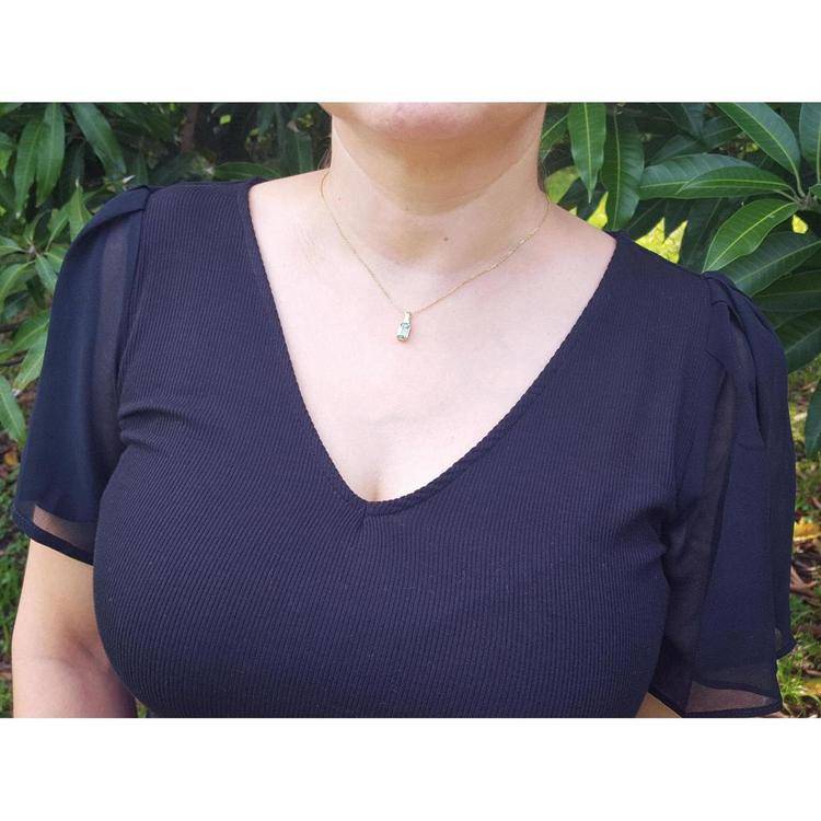A woman wearing a dark blue top with sheer sleeves and an emerald solitaire pendant against a green leafy background.