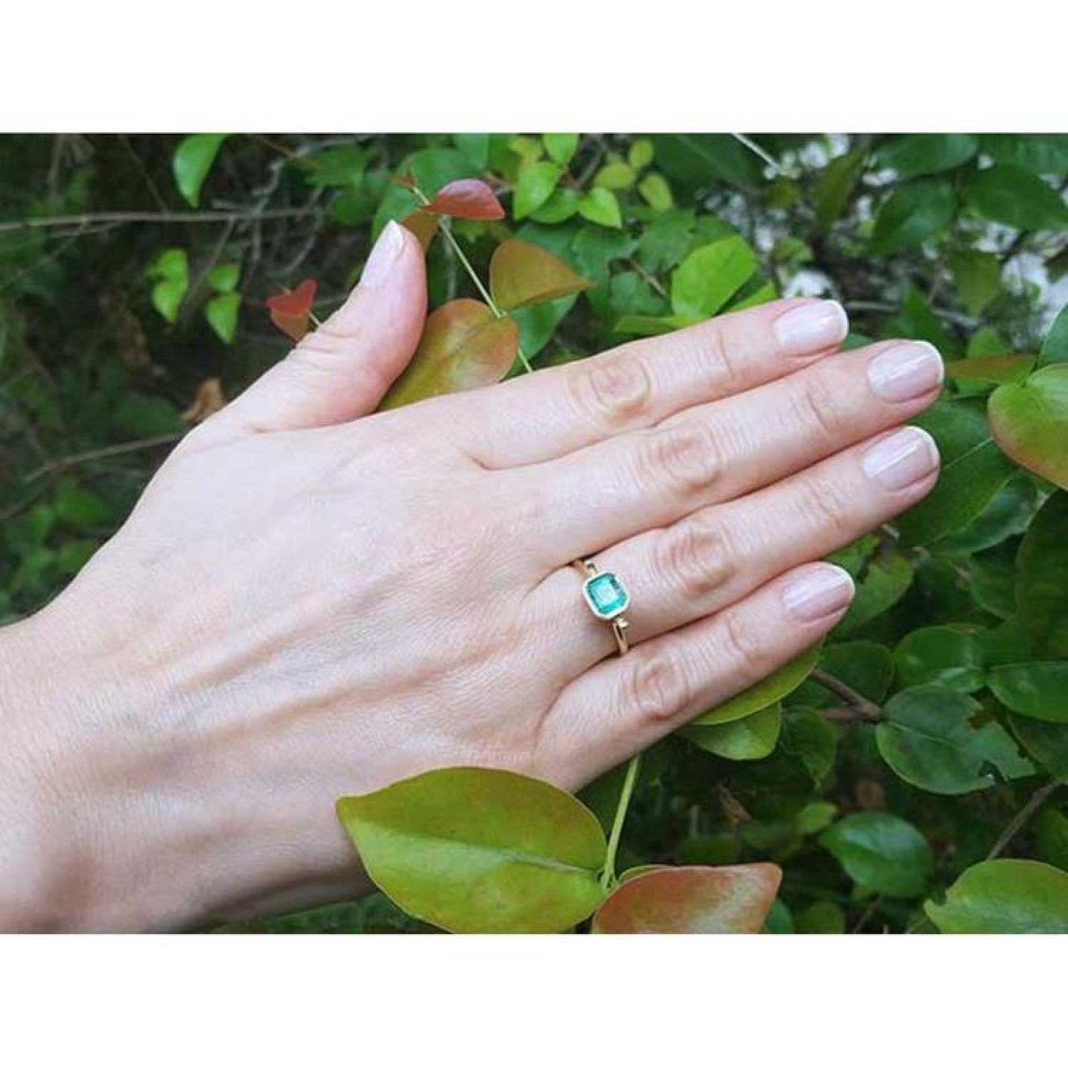 Hand wearing a 0.81 ct. medium bluish green emerald solitaire ring, bezel set in 14K yellow gold.
