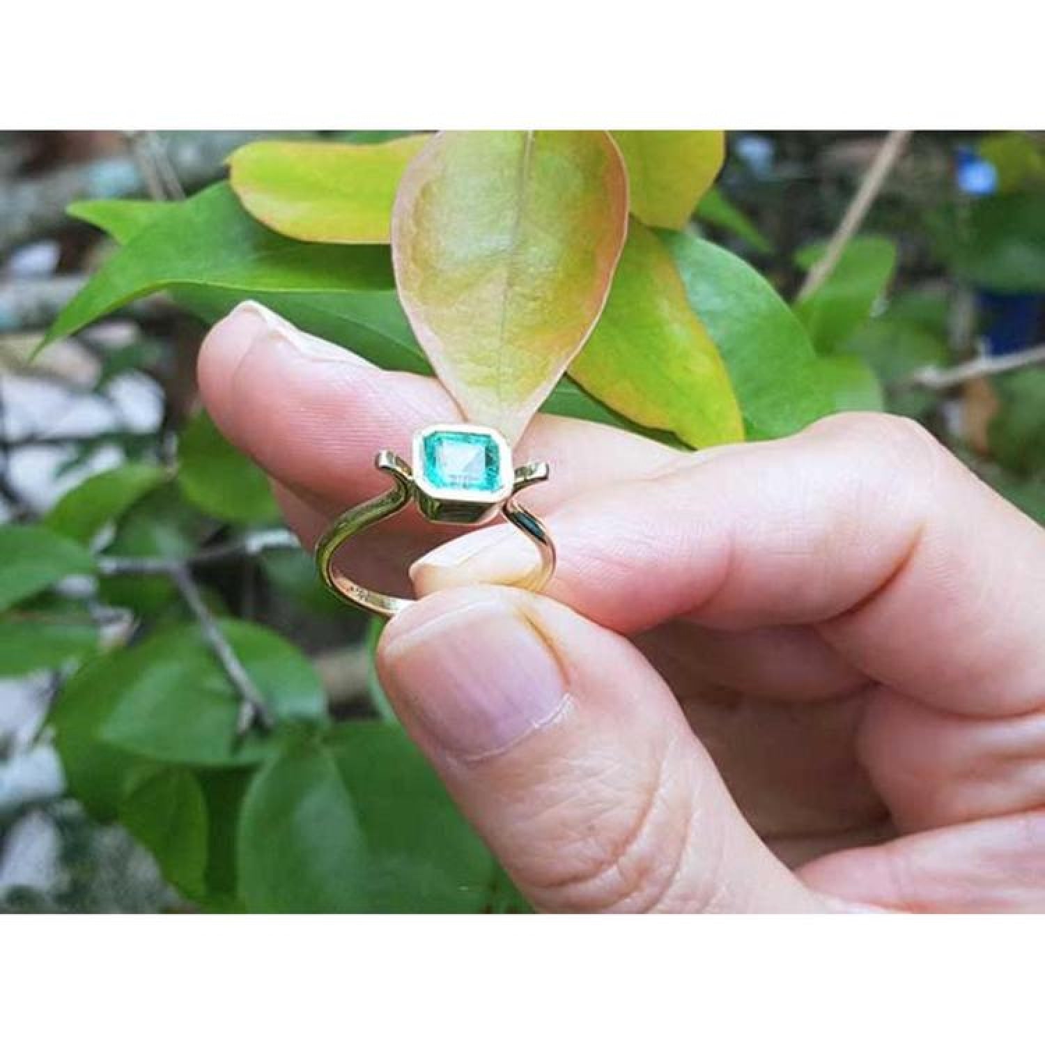 0.81 ct. emerald solitaire ring in 14K yellow gold, bezel set, held by fingers with green leaves.