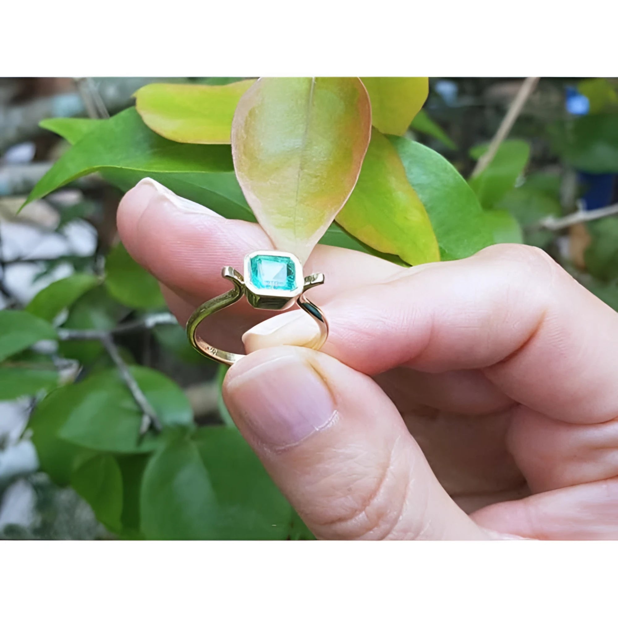 0.81 ct. emerald solitaire ring in 14K yellow gold, bezel set, held by fingers with green leaves.