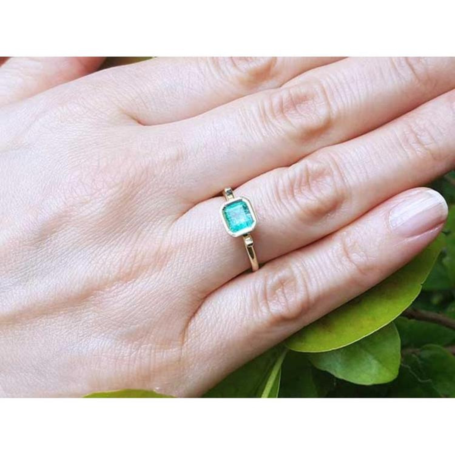 Emerald solitaire ring, bezel set in 14K yellow gold, worn on a hand with green leaves.