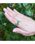 Hand wearing a mens gold ring with a natural square emerald against a green leafy background.