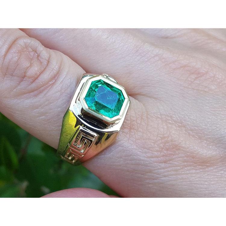 Gold ring for men with a square emerald-cut natural colombian emerald on a finger against a blurred background.