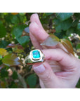 A men's gold ring with a bezel set natural emerald held by a hand against a leafy background.