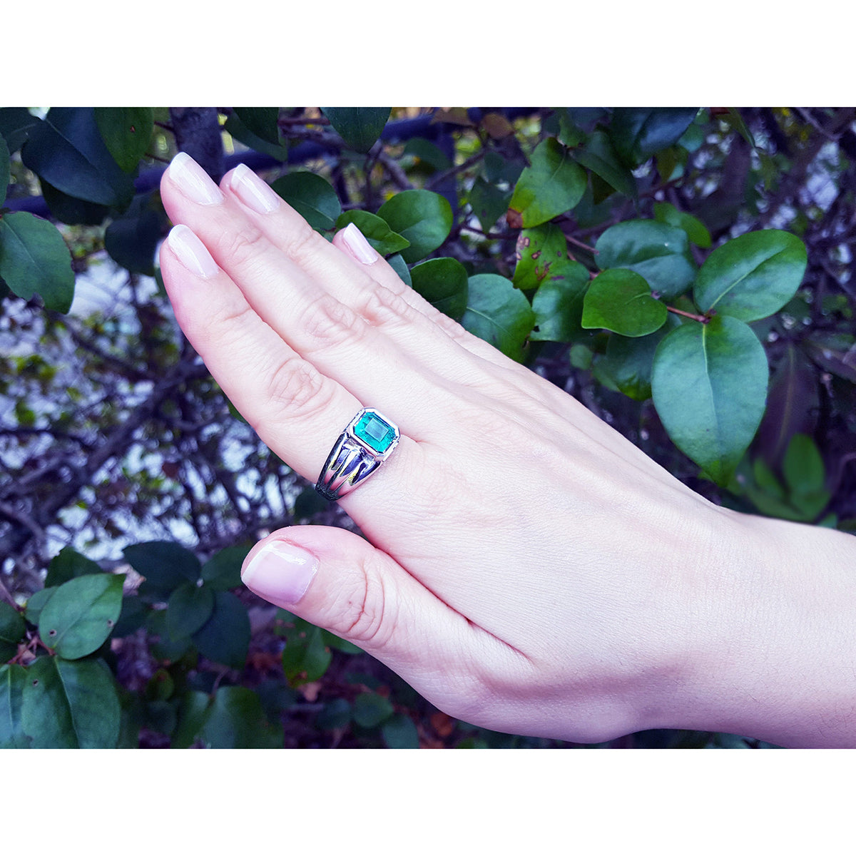 Hand wearing a ring for men with a natural emerald gemstone against a leafy background.