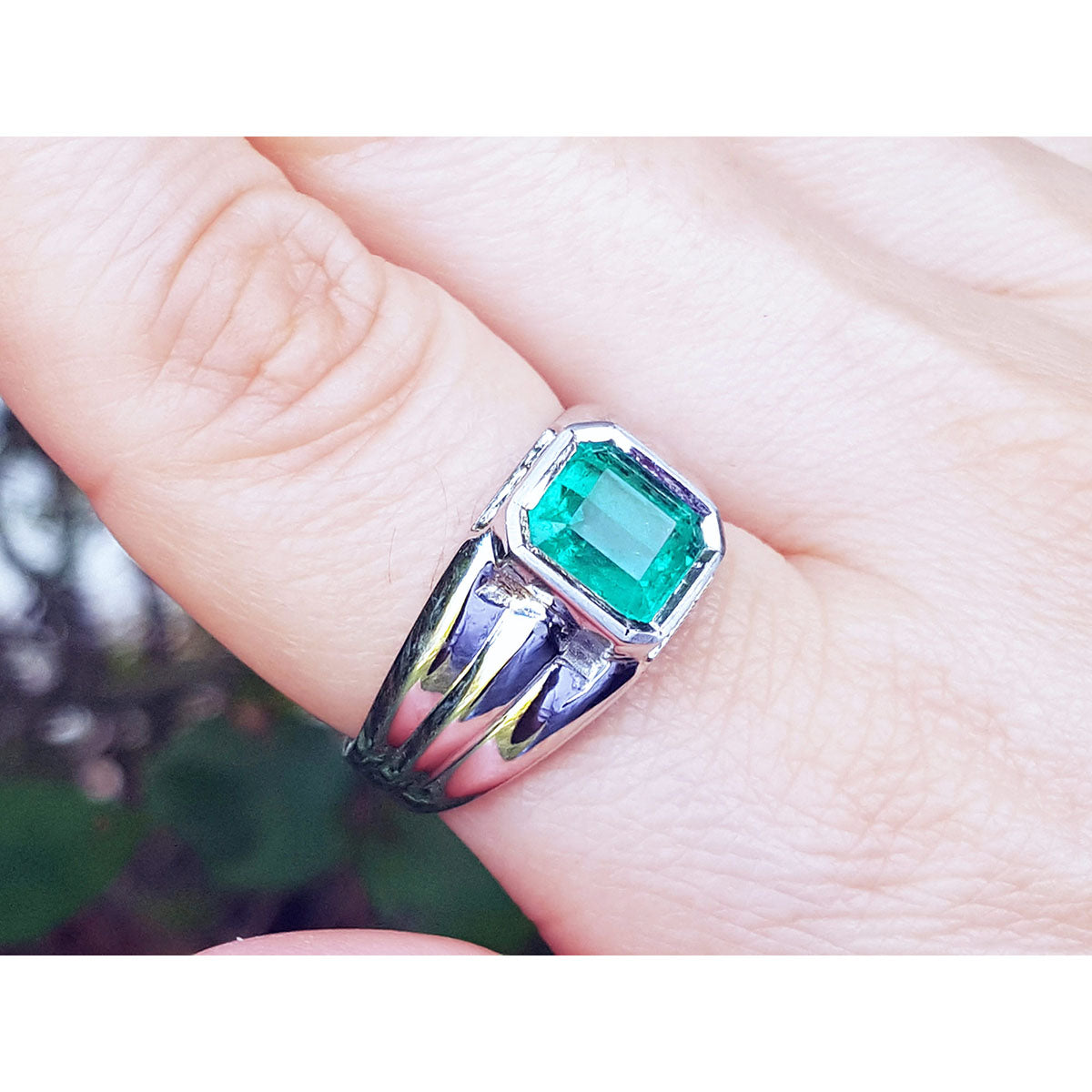 This white gold men's emerald ring is on a finger against a blurred natural background.