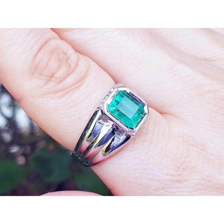 This white gold men's emerald ring is on a finger against a blurred natural background.