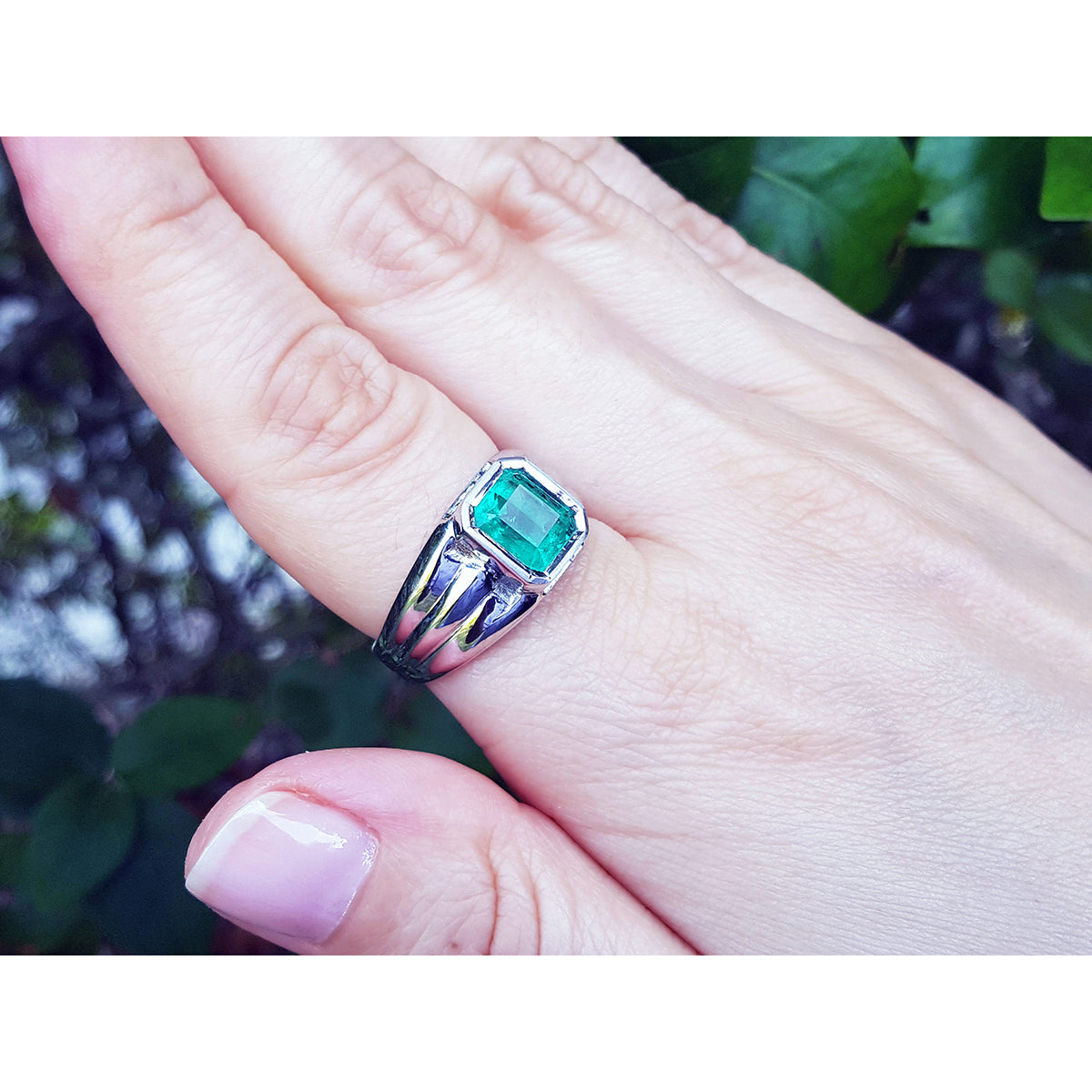 A white gold mens ring with a colombian emerald on a hand against a blurred natural background.