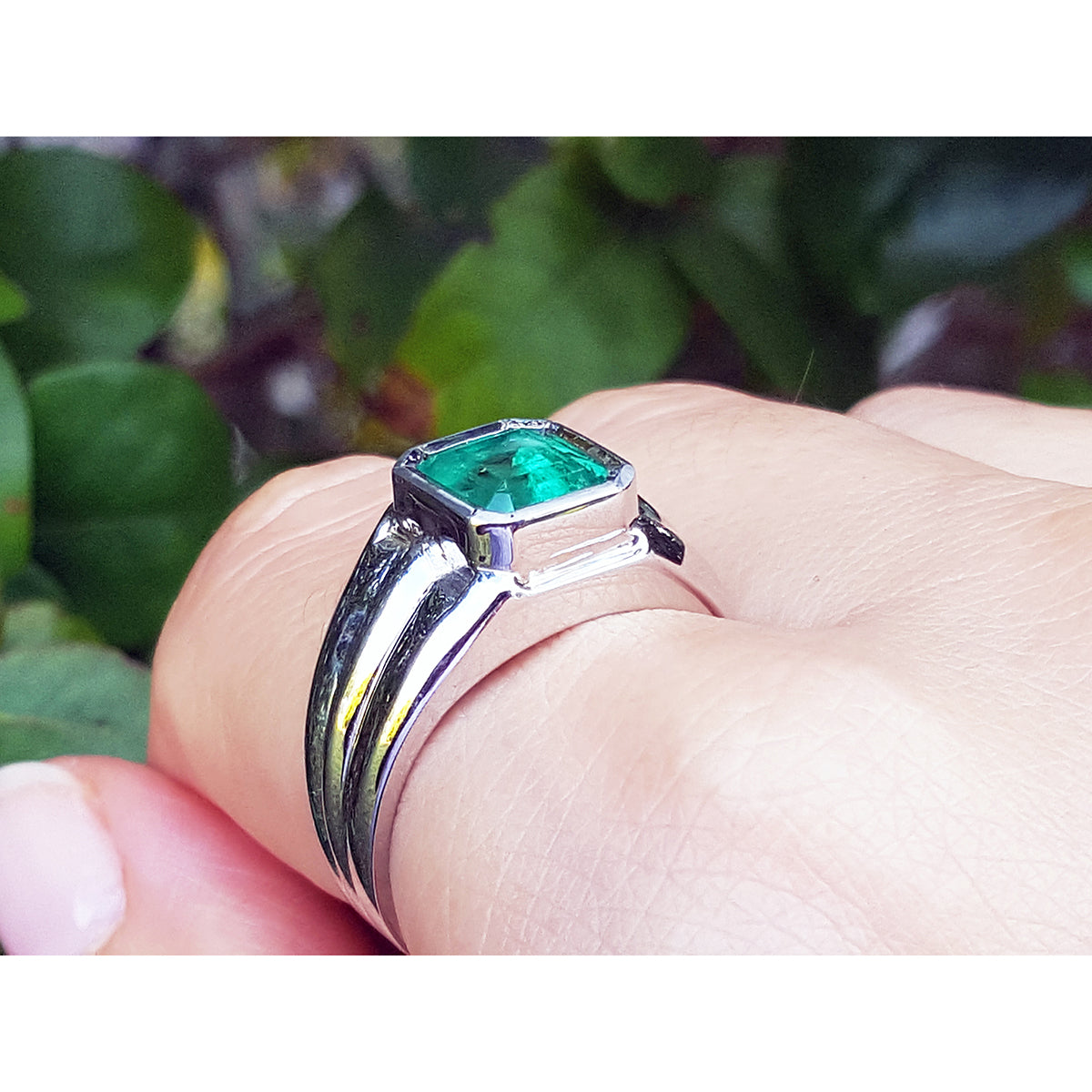 White gold mens ring with a genuine colombian emerald on a hand against a blurred green background.