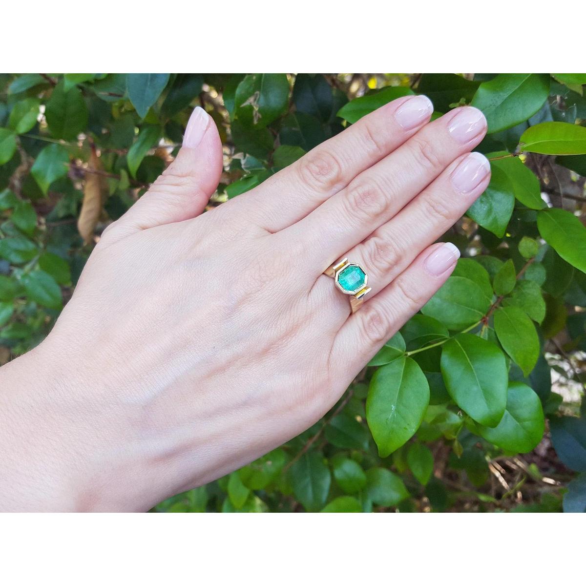 Hand wearing a gold emerald solitaire ring against a green leafy background.