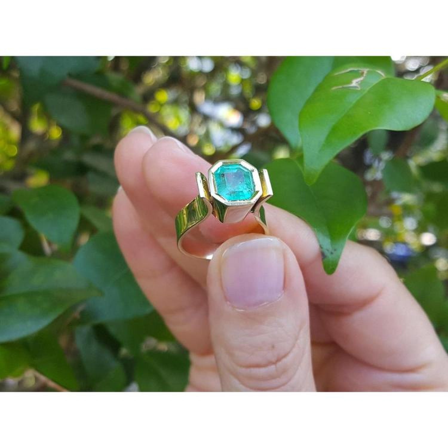 2.14 ct. rectangular emerald solitaire ring, bezel set in 14K yellow gold, held against green leaves.