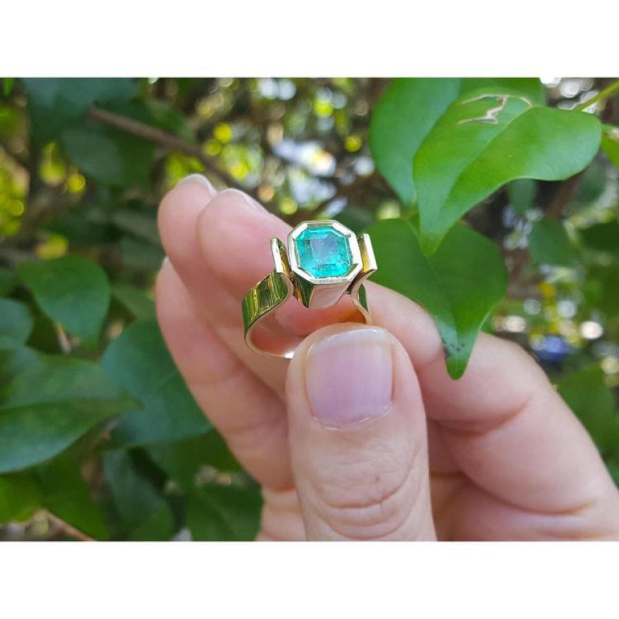 2.14 ct. rectangular emerald solitaire ring, bezel set in 14K yellow gold, held against green leaves.