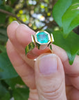 A Colombian emerald solitaire ring for women held by a hand against a green leafy background.