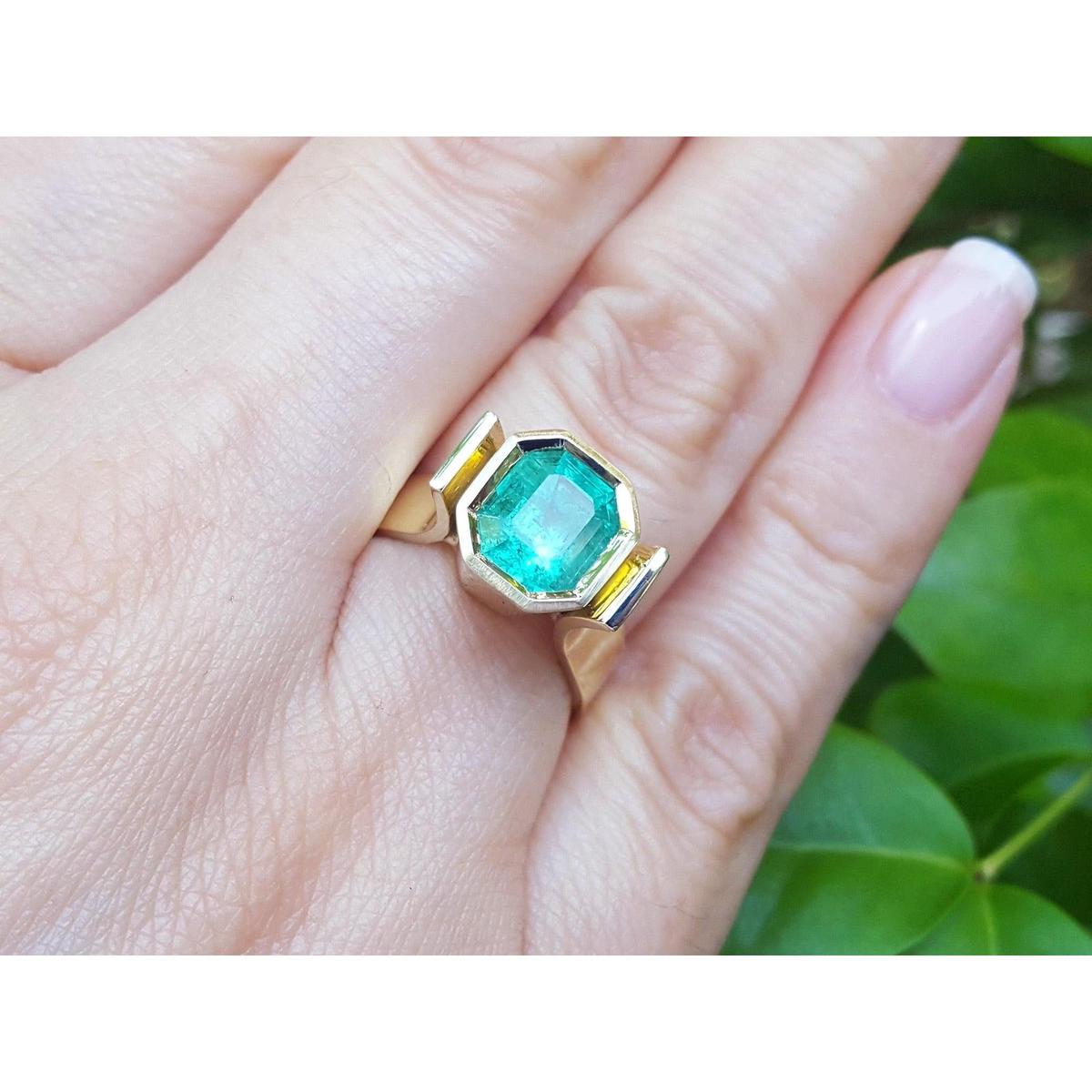 A women's emerald solitaire ring bezel set on a hand against a green leafy background.