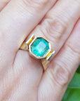 A women's emerald solitaire ring bezel set on a hand against a green leafy background.