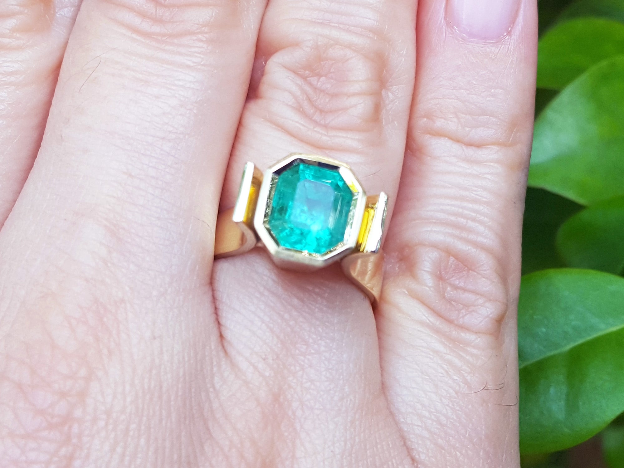 Emerald solitaire ring yellow gold on a hand, with green leaves in the background