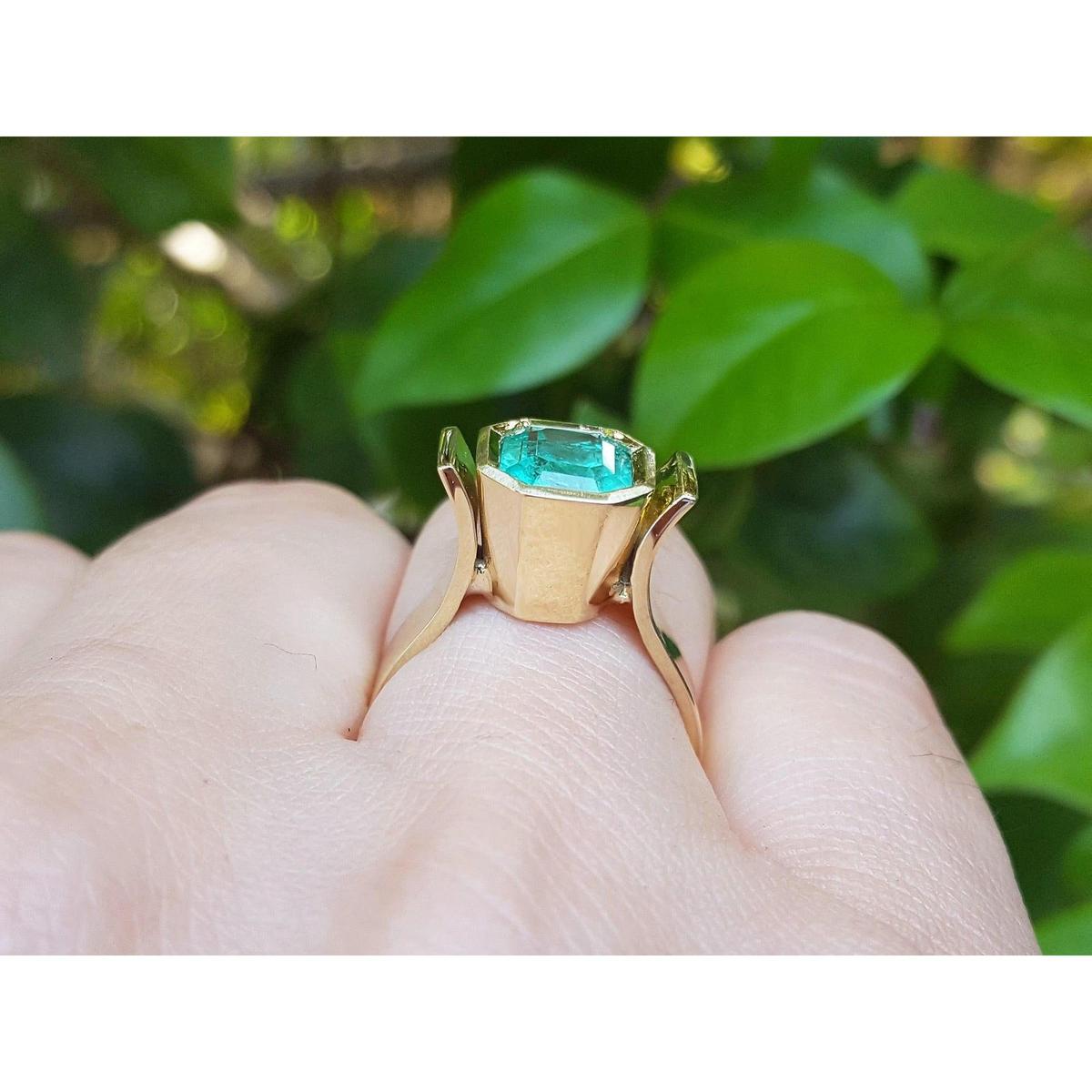 A bezel set emerald solitaire ring in yellow gold on a person's finger against a green leafy background.