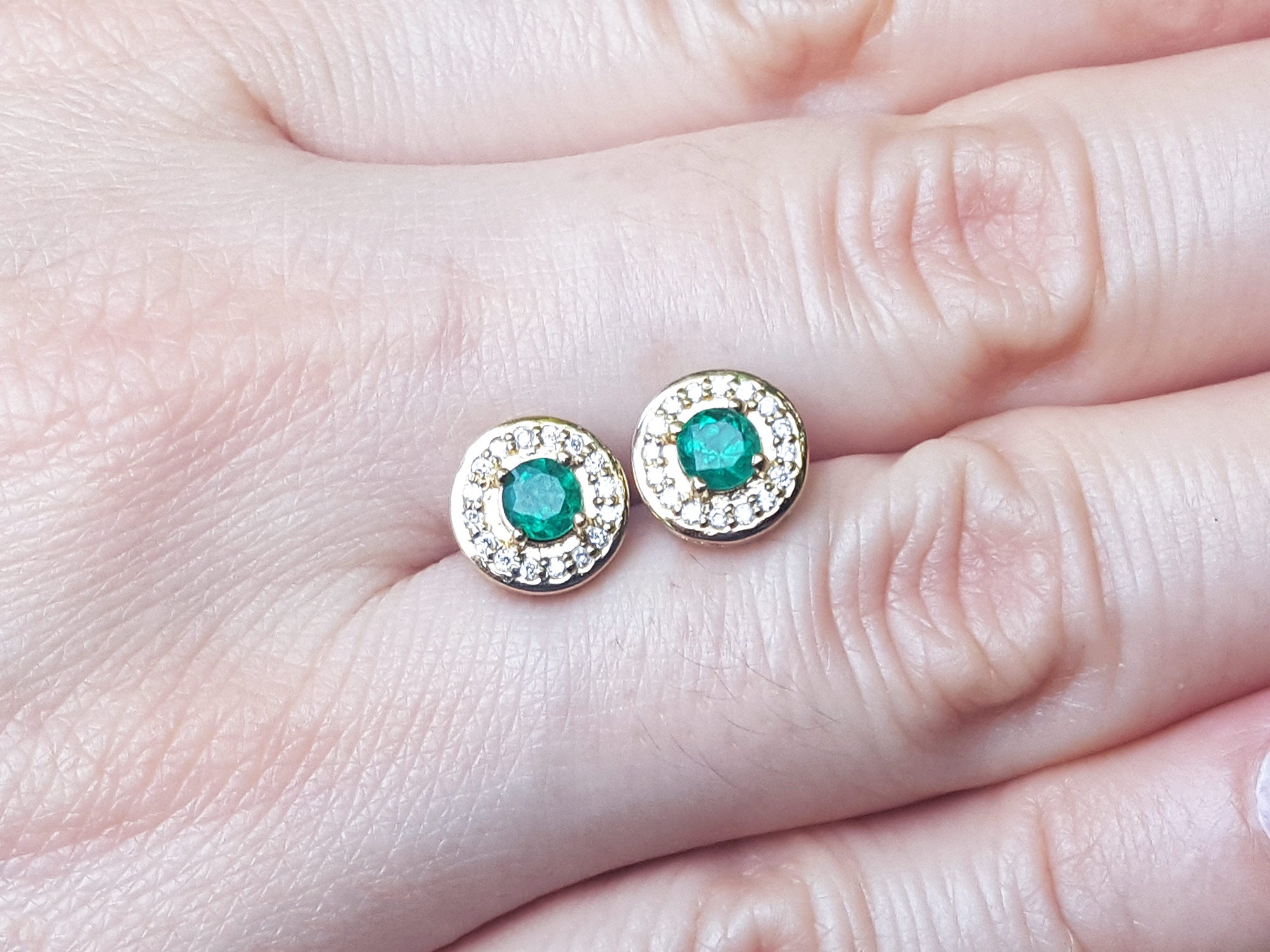 Gold earrings with emerald gemstones held between fingers on a light skin background.