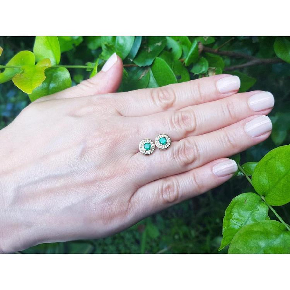 14k yellow gold emerald halo diamond stud earrings on a hand, deep bluish-green round emeralds.
