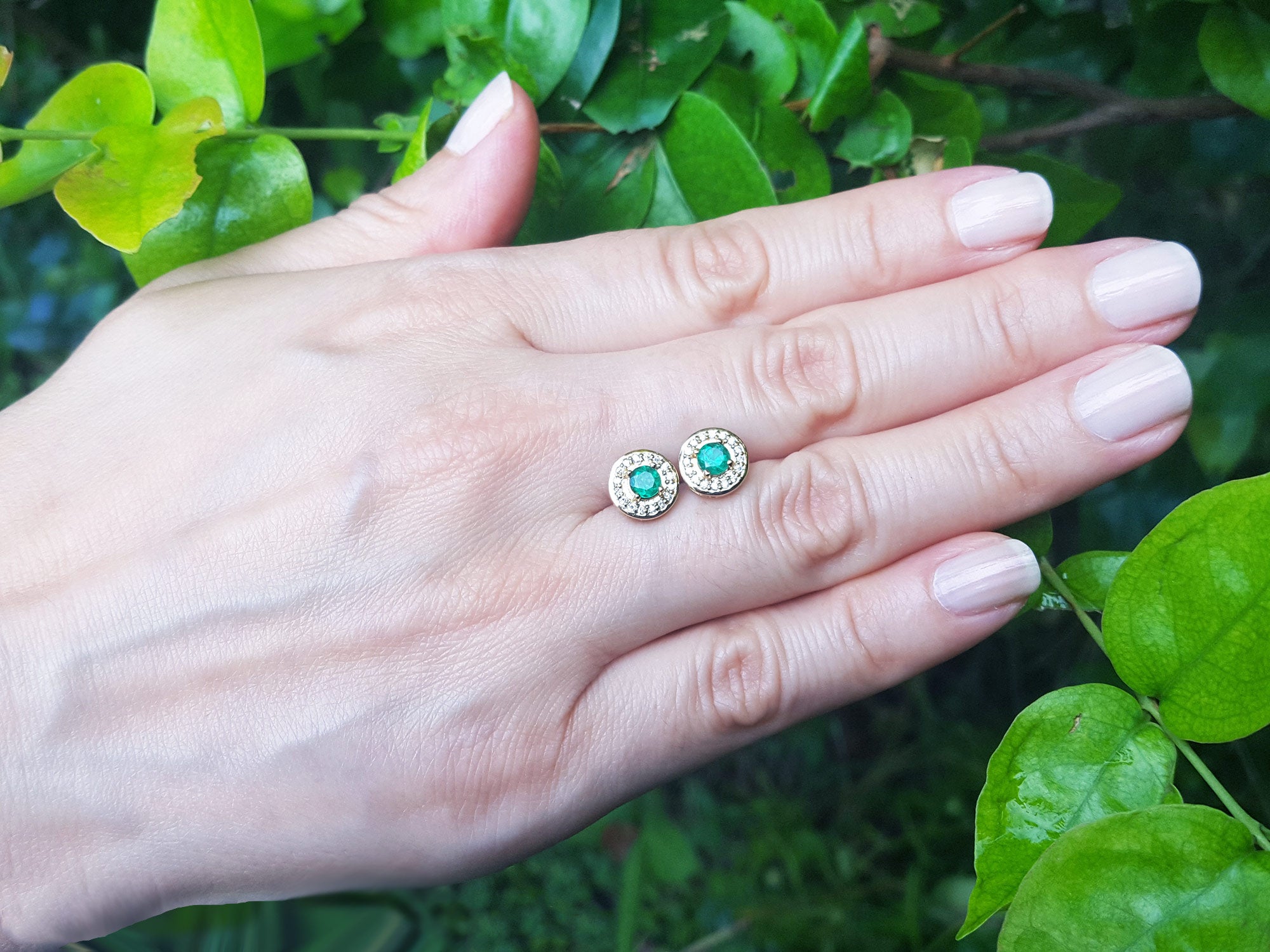 Hand holding a pair of emerald earrings with a green leafy background.