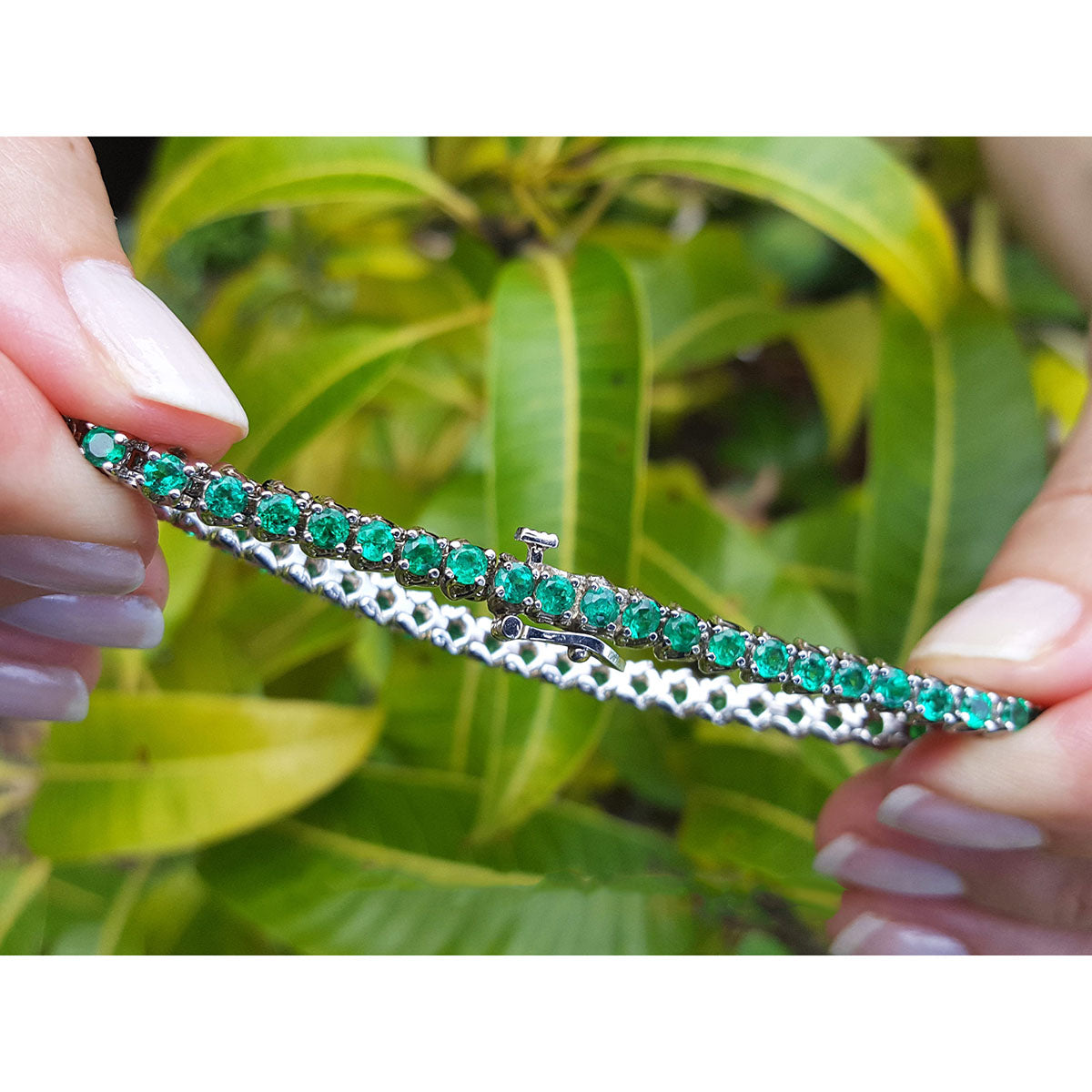 Natural emerald tennis bracelet held by a hand between fingers with a green leafy background.