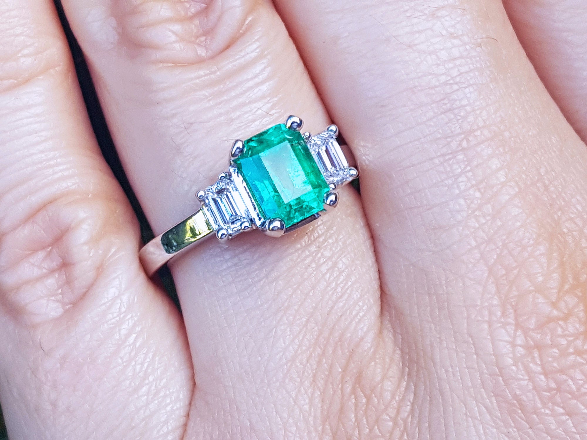 Emerald ring with emerald-cit side diamonds on a finger.