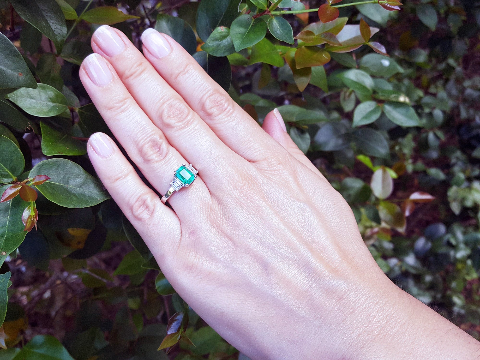 Hand wearing a natural emerald and side diamonds ring for women against a leafy background.