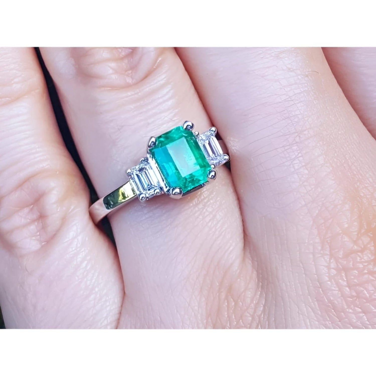 18K white gold three-stone emerald ring with a central bluish-green emerald and side emerald-cut diamonds.