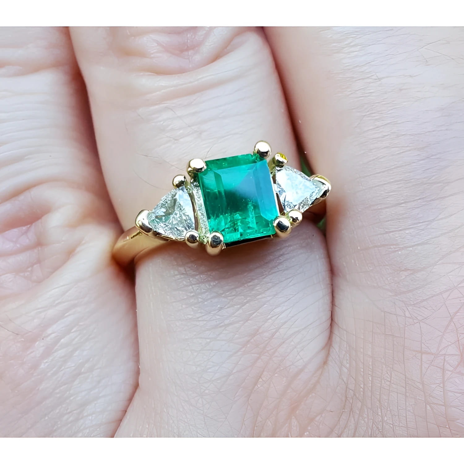 A yellow gold natural colombian emerald ring with side genuine trillion cut diamonds on a finger.