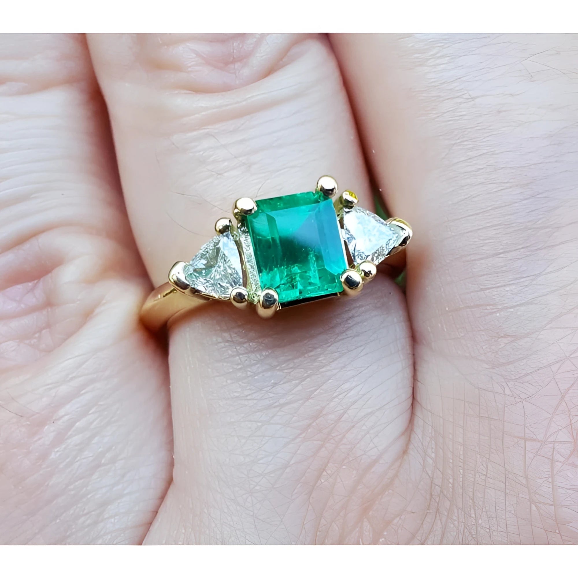 A yellow gold natural colombian emerald ring with side genuine trillion cut diamonds on a finger.