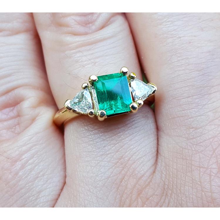 A yellow gold natural colombian emerald ring with side genuine trillion cut diamonds on a finger.
