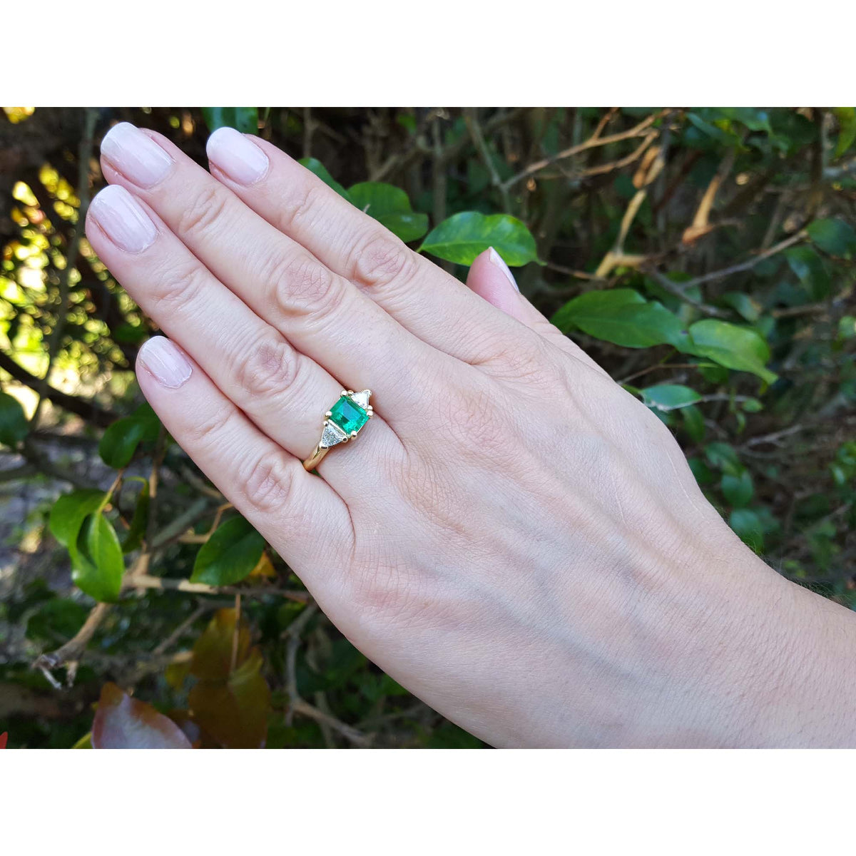 Hand wearing a three stone colombian emerald and trillion side diamond ring against a natural background.
