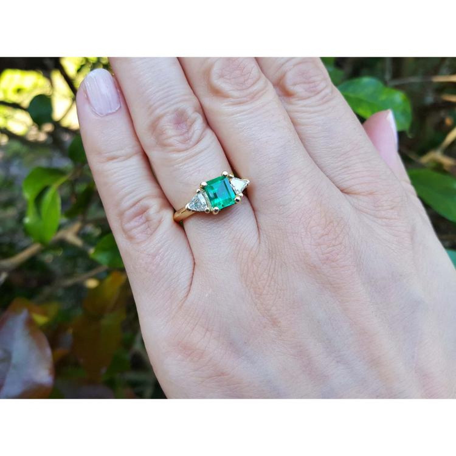 1.48 ct. emerald and trillion side diamond ring in 18K gold on a hand.