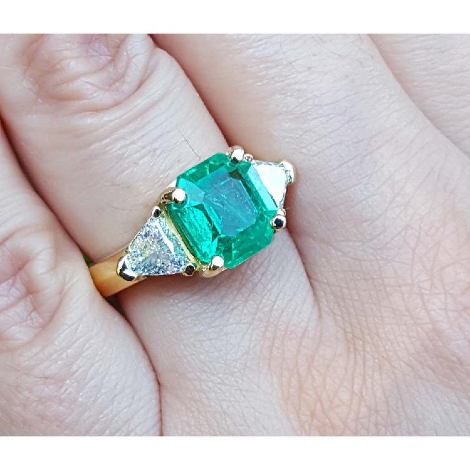 18k yellow gold ring with a large green emerald and two trillion side diamonds on a finger.