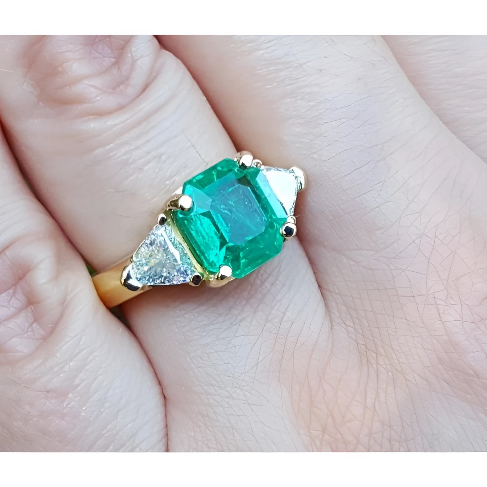18k yellow gold ring with a large green emerald and two trillion side diamonds on a finger.