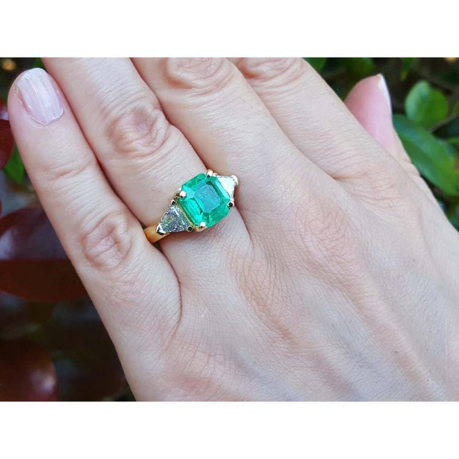 18k yellow gold ring with a large green emerald and two trillion side diamonds on a hand.