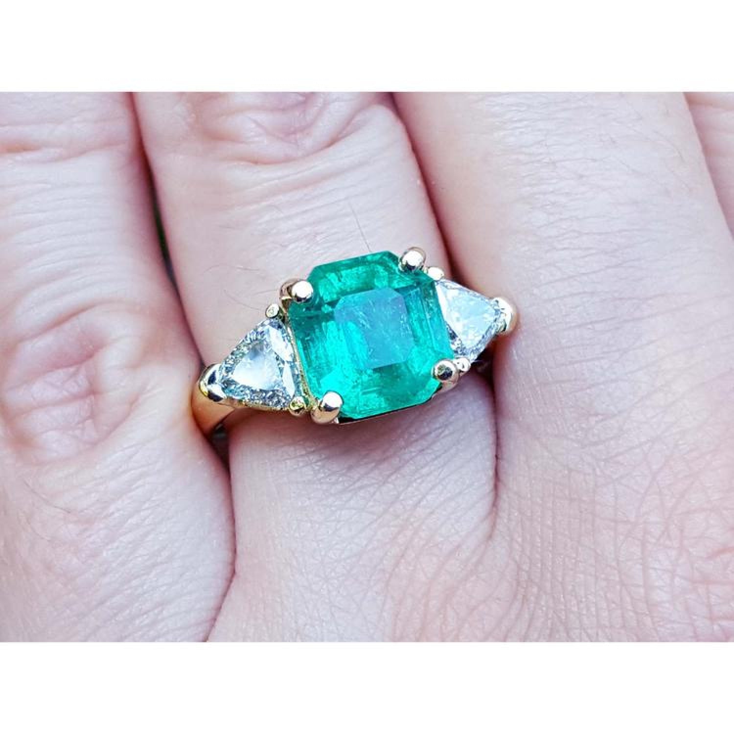 18k yellow gold ring with a large green emerald and two trillion side diamonds on a finger.